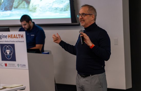 A speaker presenting near a screen, gesturing with hands.