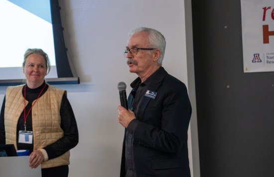 Two women standing near a wall, one of them presenting.