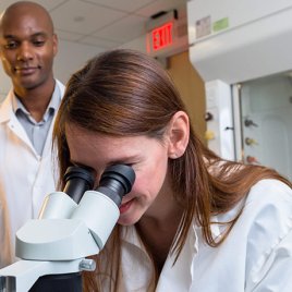 Dr. Deveroux Ferguson Observes a Lab Associate as She Looks through a Microscope